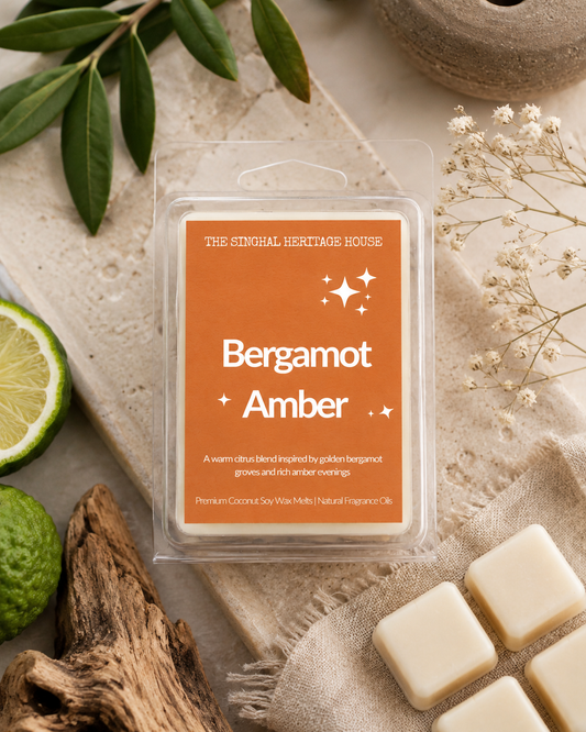 A clear plastic clamshell package labeled 'Bergamot Amber' by The Sizzling Heritage House, shown on a styled flat-lay with a beige fabric surface, green leaves, a lime wedge, dried flowers, and cream-colored wax cubes.